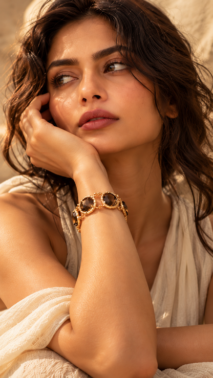 A woman in a cream draped garment wearing a gold-toned bracelet with dark oval stones, posed in warm, soft lighting.