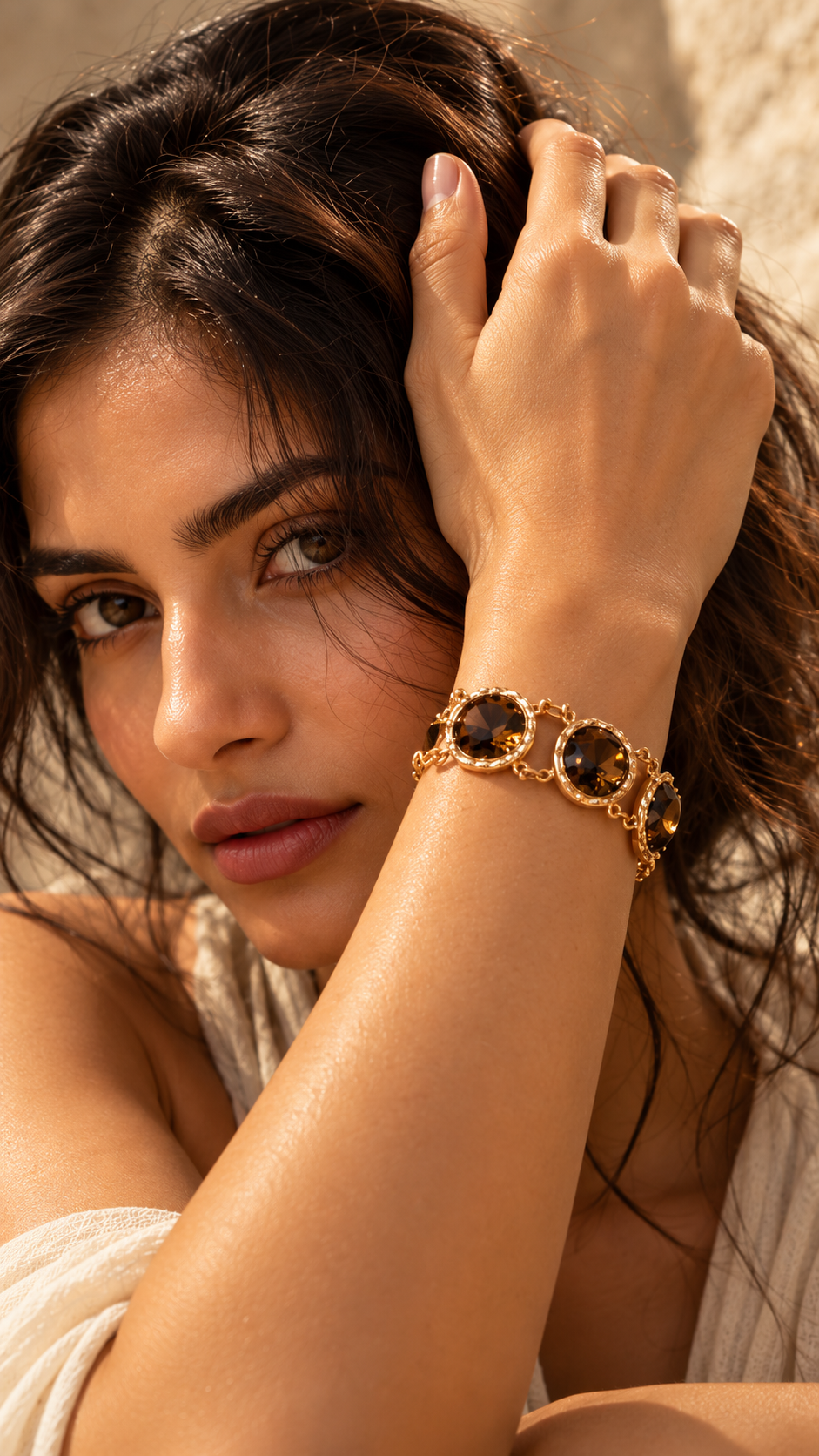 A close-up lifestyle shot of a woman wearing a gold-toned bracelet with three round dark brown stones, set against a warm, sunlit neutral background.
