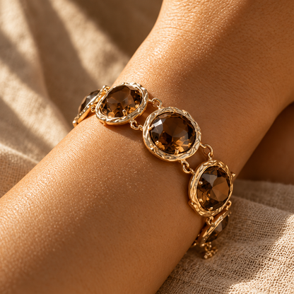 A gold-toned bracelet with three large oval brown faceted gemstones, shown worn on a person's wrist against a textured beige fabric background.