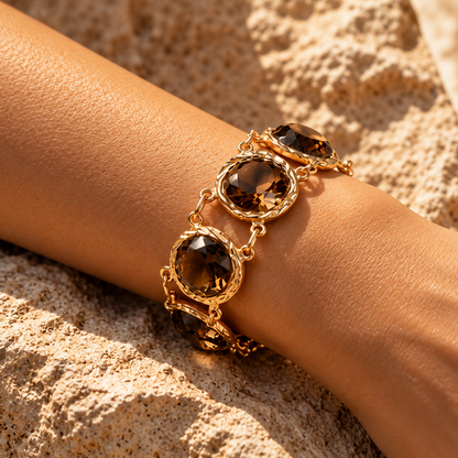 A gold-toned bracelet with three large faceted smoky brown gemstones, shown worn on a person's wrist against a sandy, sunlit background.