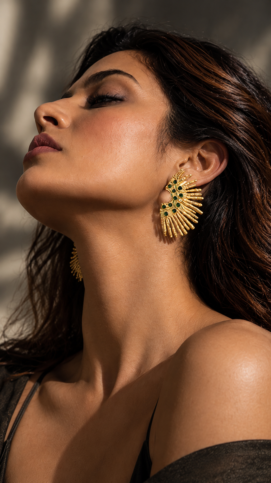 Side-profile view of a model wearing Medoso Vimosa sculptural statement earrings with gold rays and deep green stones.