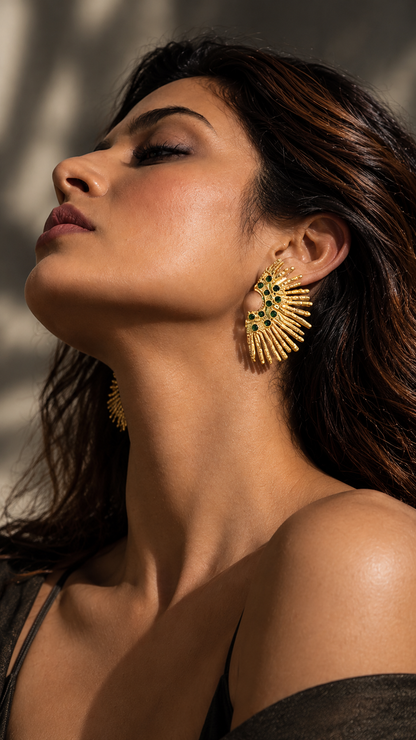 Side-profile view of a model wearing Medoso Vimosa sculptural statement earrings with gold rays and deep green stones.